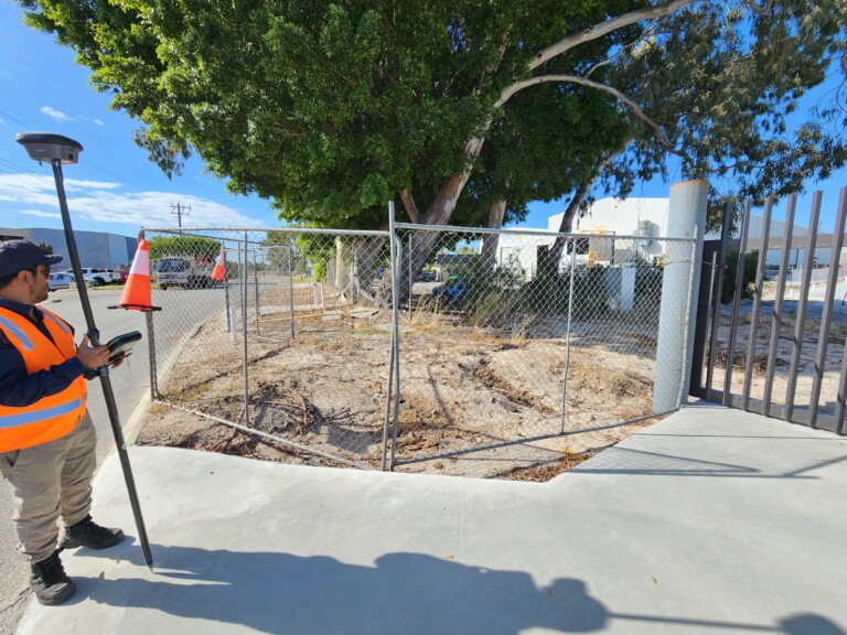 A surveyor in high-visibility orange vest holds a GPS survey pole and tablet on a concrete footpath beside a chain-link temporary construction fence enclosing a large eucalyptus tree on a vacant industrial lot.