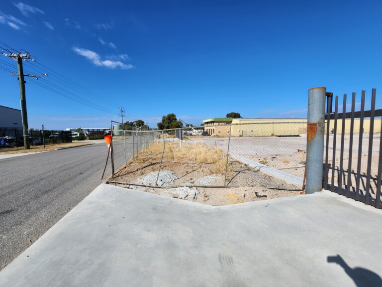 View along a concrete footpath beside a vacant, sandy industrial lot enclosed by temporary chain-link fencing.