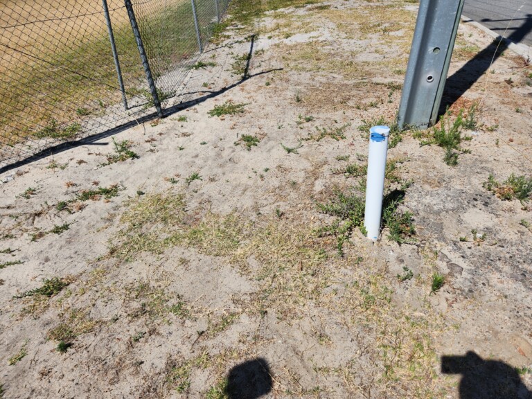 A single white PVC monitoring well casing with a blue cap installed in sandy ground beside a galvanised steel fence post, with sparse weeds and a chain-link fence visible in the background.