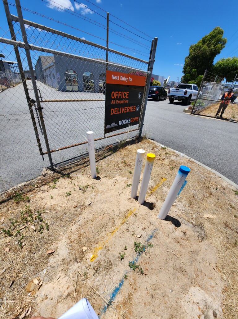 Four white PVC monitoring well casings — one with a white cap, two with yellow caps, and one with a blue cap — installed in sandy ground near a chain-link gate bearing a "RocksGone" business sign directing visitors to the office and deliveries entrance.