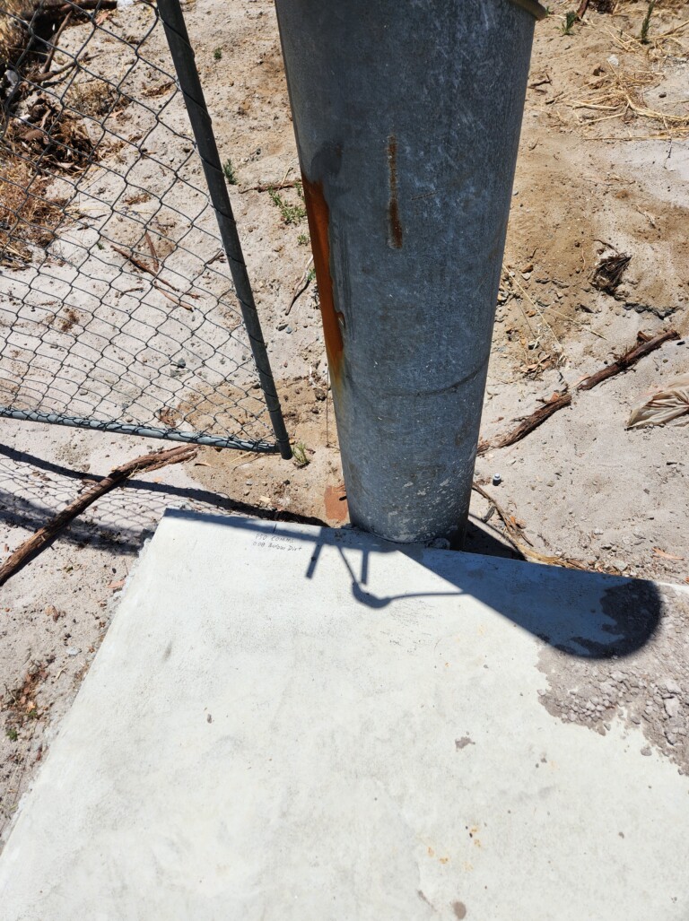 Close-up of the base of a large rusted galvanised steel gate post set into a concrete pad, with a handwritten notation reading "PSO Comms 0.08 Below Dirt" on the concrete surface. Sandy ground and chain-link fencing are visible alongside.