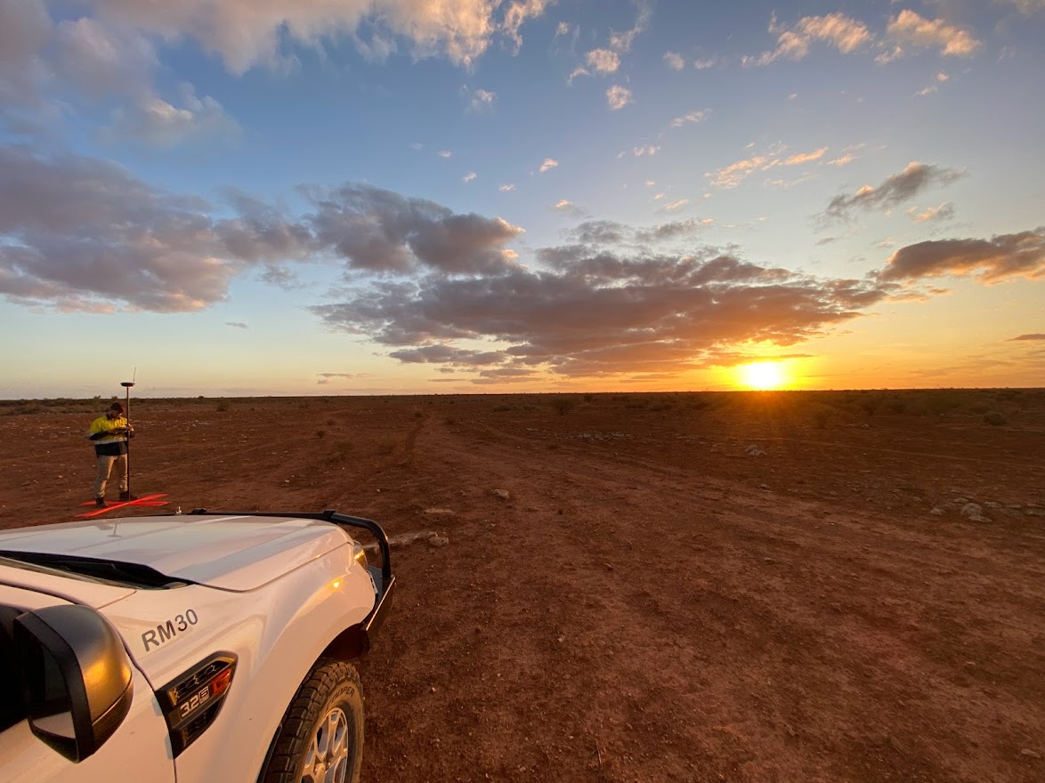 Topographic surveyor conducting field measurements at sunset in a remote, open landscape beside a survey vehicle.