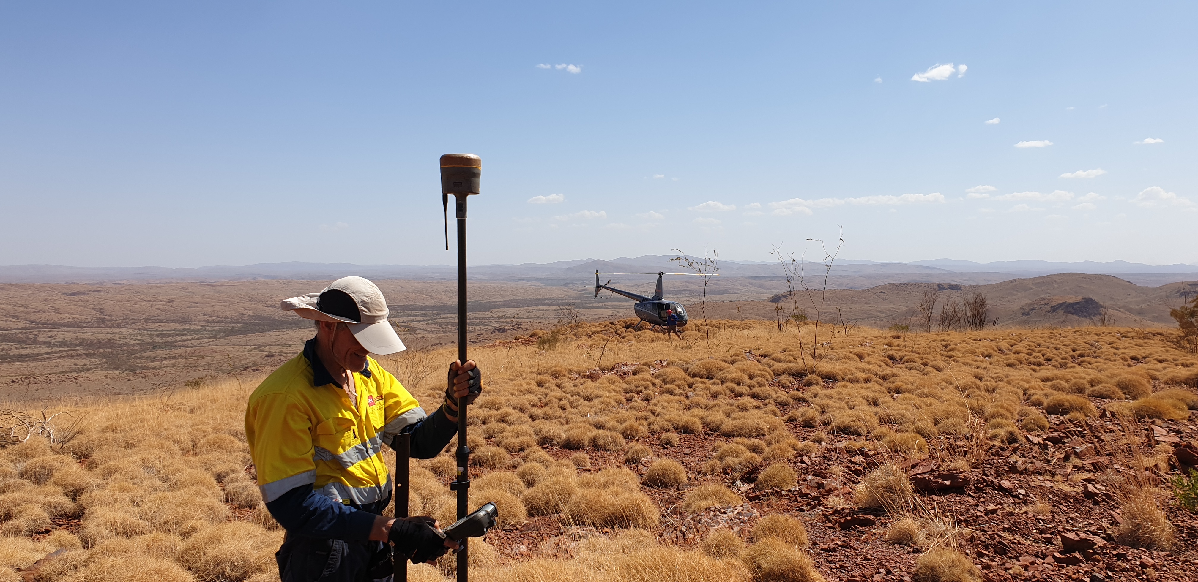 Topographic surveyor using GPS equipment in remote terrain, with a helicopter landing in the background.